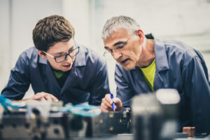 An older man, practicing senior mentoring, points out the parts of a piece of equipment to a young man.