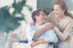 An older woman smiles in her wheelchair, knowing she can age comfortably at home with the help of her caregiver.