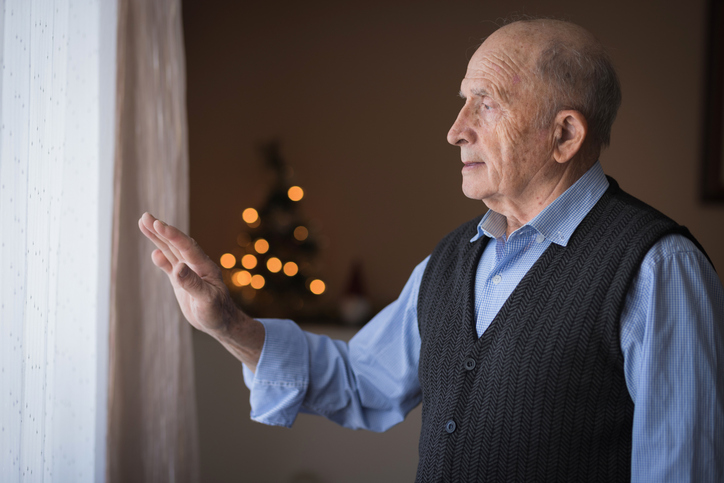 An older man struggling with seasonal affective disorder gazes out the window and offers a halfhearted wave .