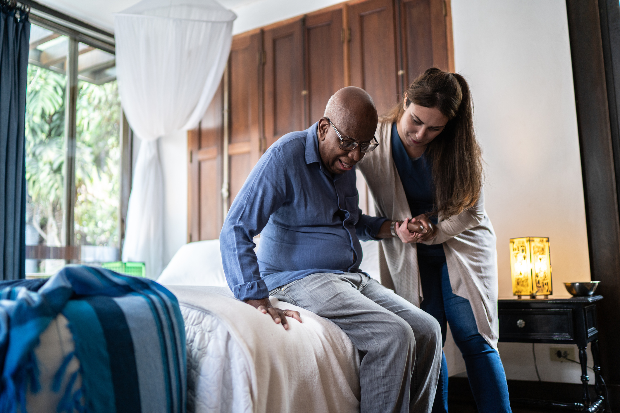 An older man receives assistance to get out of bed with the help of his live-in caregiver.