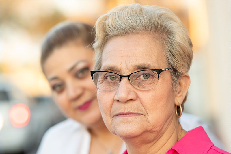 An older woman looks stoically ahead while a younger woman, who knows what caregivers say they need, stands blurred behind her.