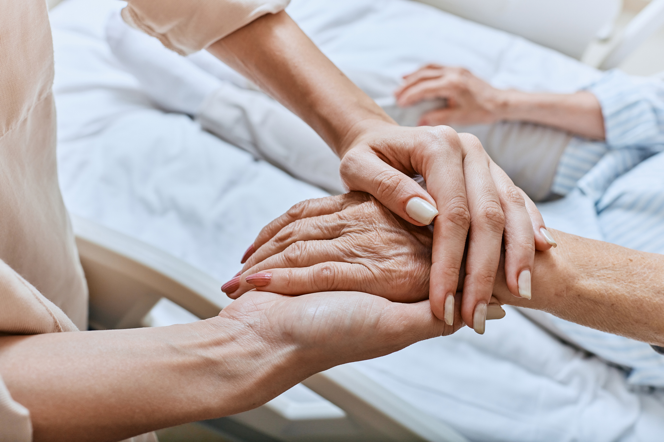 A younger woman’s hands hold the hand of an older loved one experiencing end-stage dementia.