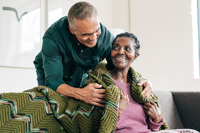 A senior man covers his wife with a cozy blanket as he strives to avoid common Alzheimer’s caregiver mistakes.